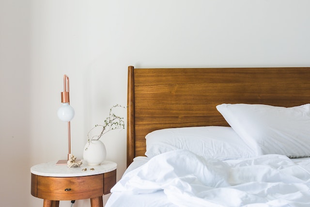 bed with a wooden frame and white sheets next to a night table with a lamp and a small plant on it